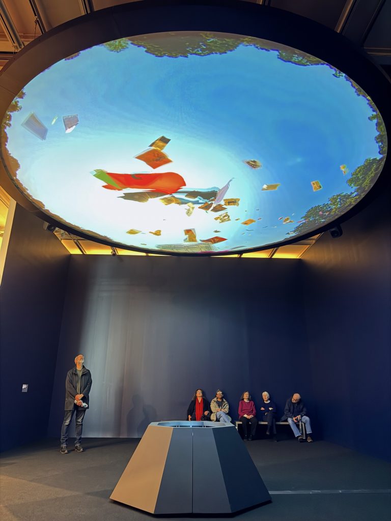 An installation view of Michelle Lopez’s work at the Whitney Biennial, showing several visitors sitting beneath a large, circular ceiling projection of a clear blue sky.