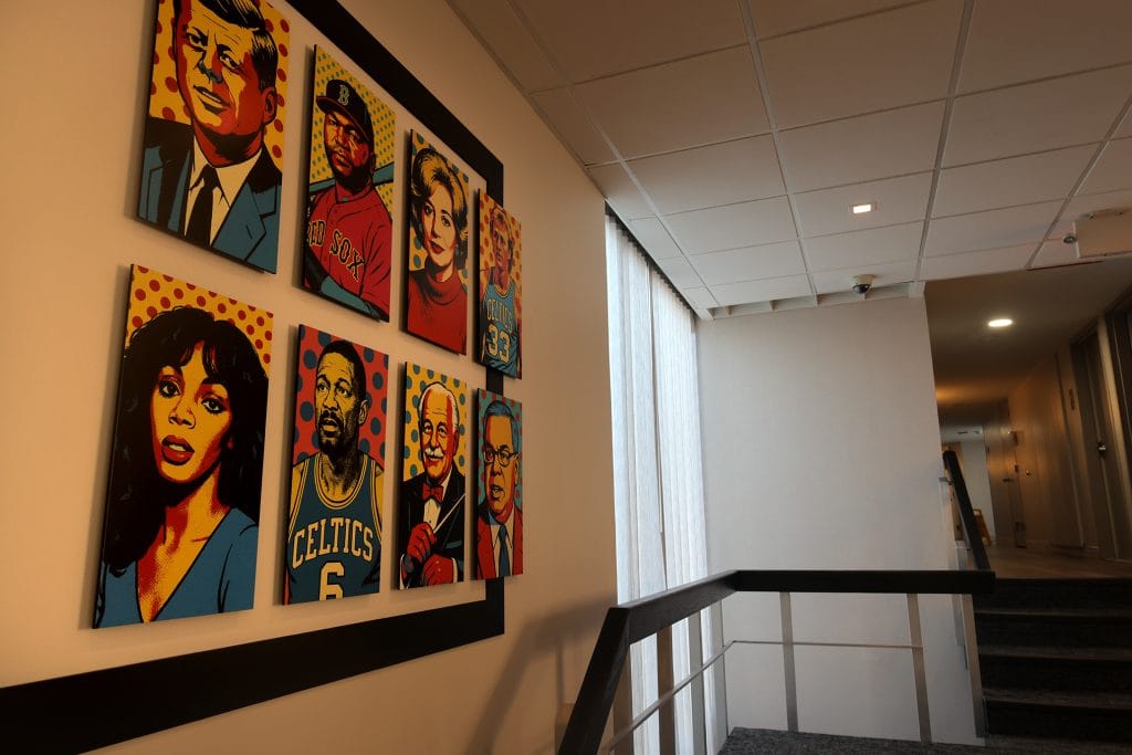 A photo of a hotel stairwell with a wall-mounted grid of colorful pop-art style portrait paintings depicting adult men and women of varied races and ages, rendered in bold colors and halftone dots, with a modern railing and ceiling lights visible to the right.
