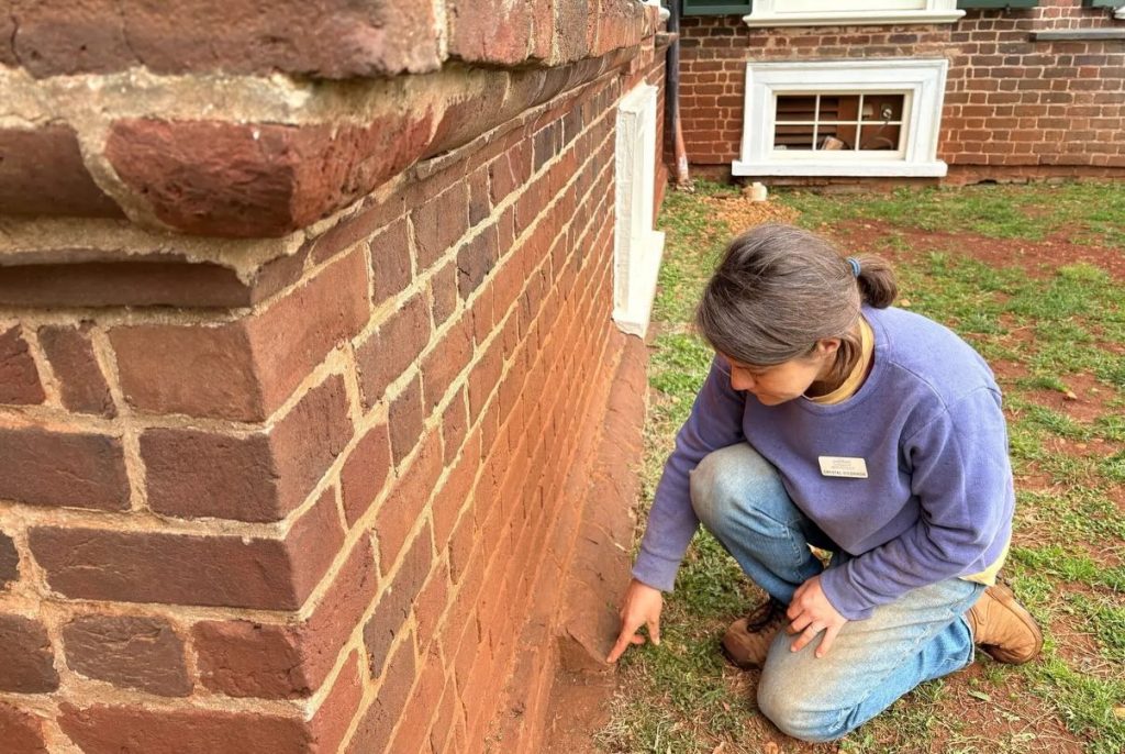 A photograph of an archaeologist kneeling and pointing to curved bricks at the base of where a brick wall meets grass