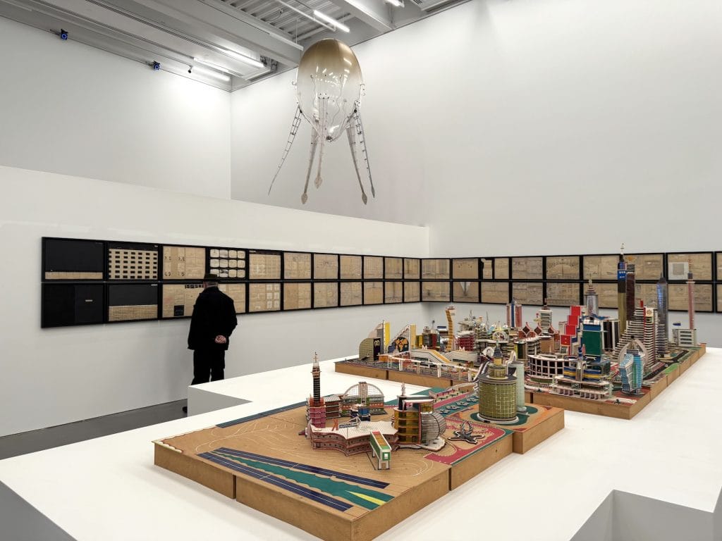 A vast gallery features a dense architectural model city on white platforms, a row of framed drawings, and a jellyfish-like sculpture suspended from the ceiling.