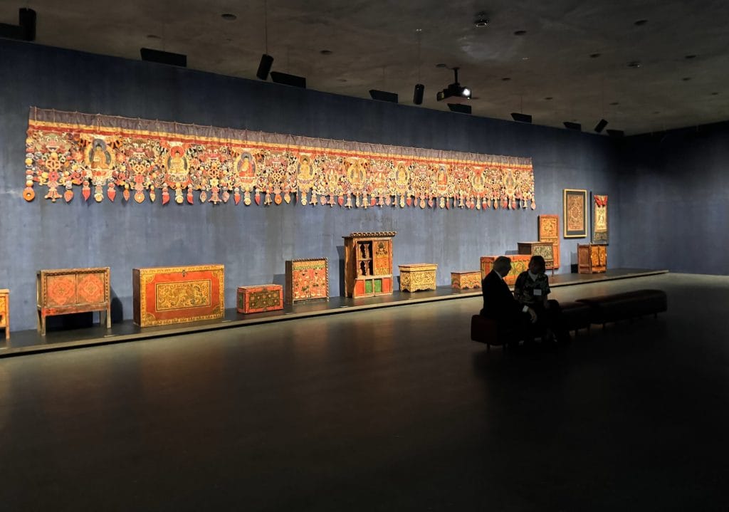 A long, ornate Buddhist textile hangs above several intricately painted wooden chests in a blue-walled gallery. Two visitors sit on a bench observing the display.