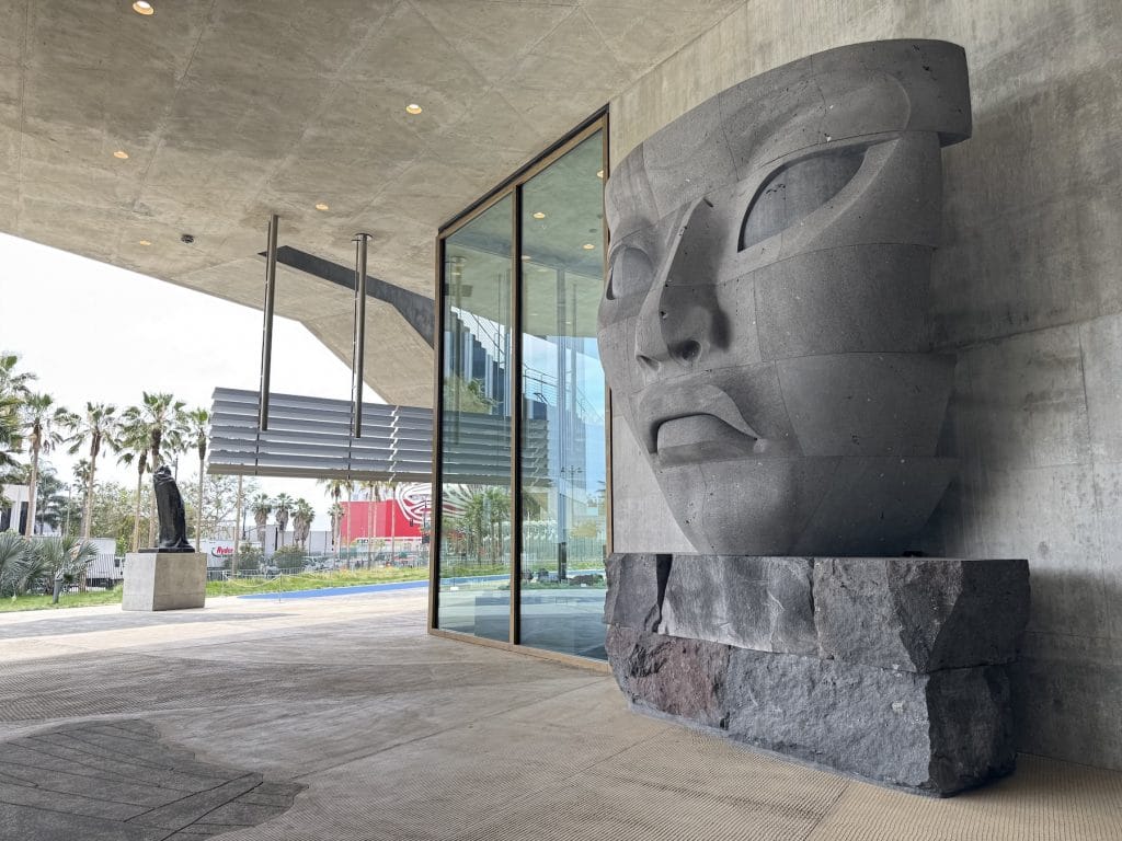 A massive, volcanic stone head sculpture rests on a raw rock base outside a concrete museum building, featuring stylized, ancient-inspired features and stacked horizontal segments.