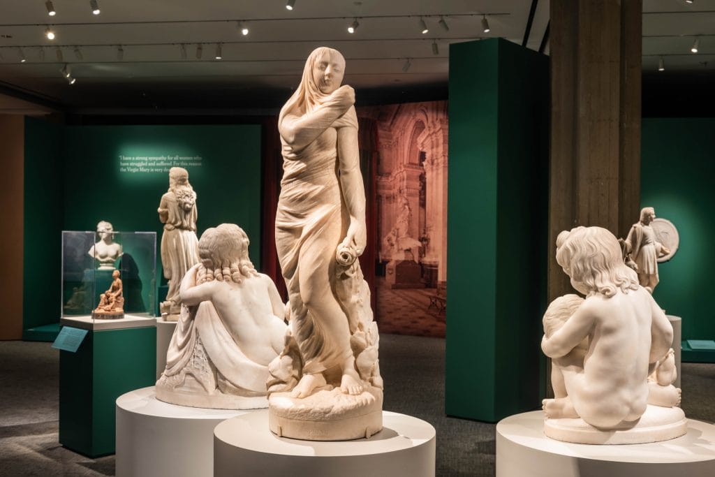A photo of a museum gallery displaying several white marble sculptures on pedestals, including a life-size carved woman draped in fabric at center and smaller figures of children and classical subjects around her, under soft overhead lighting.