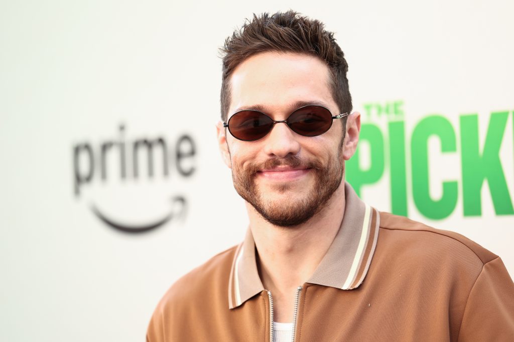 A photograph of Pete Davidson from the chest up, wearing a tan shirt jacket and ovular brown sunglasses