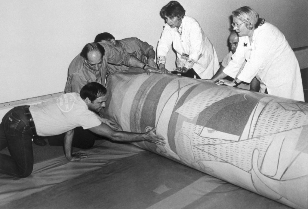 Handlers carefully roll massive canvas painting across floor, guiding cylindrical bundle with coordinated effort indoors