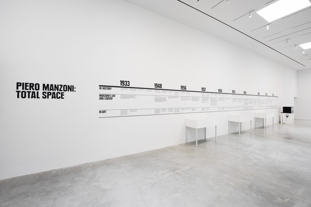 Minimal white gallery interior with wall text timeline titled Piero Manzoni Total Space and sparse display tables