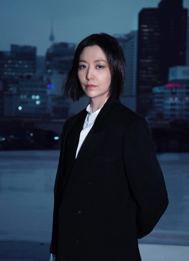 Portrait of an East Asian woman in shoulder length dark hair, sporting black blazer and white shirt, posing against backdrop of skyscrapers.