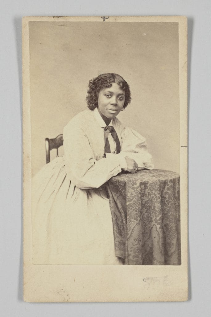 A black and white photo of a young Black woman, likely in her 20s or 30s, with short curly hair, seated beside a table draped with patterned fabric, wearing a light-colored dress with a dark ribbon at the collar, and looking calmly toward the camera.