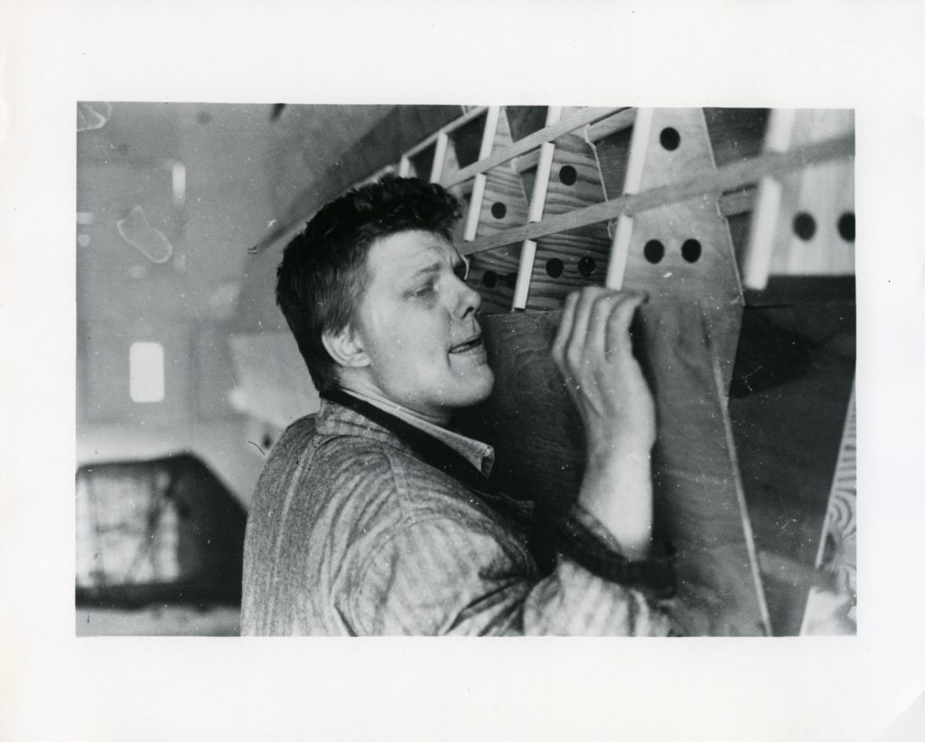 Black-and-white photo of a man lifting and steadying a large wooden structure indoors, seen in profile.