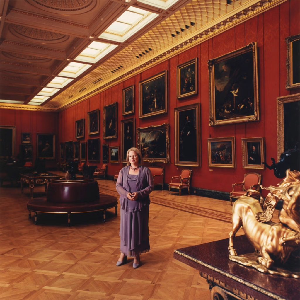 A photo of an older white woman with short blonde hair standing in an opulent museum gallery. She wears a flowing lavender dress with a matching jacket and shoes. She stands on a polished parquet floor surrounded by red walls densely hung with large framed paintings, ornate gold accents, upholstered chairs, and a sculptural gilded centerpiece in the foreground.