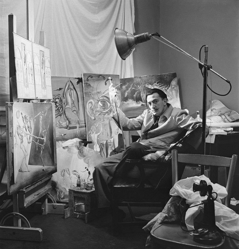 Salvador Dali seated in studio surrounded by surreal paintings, holding brush, beneath large adjustable lamp indoors