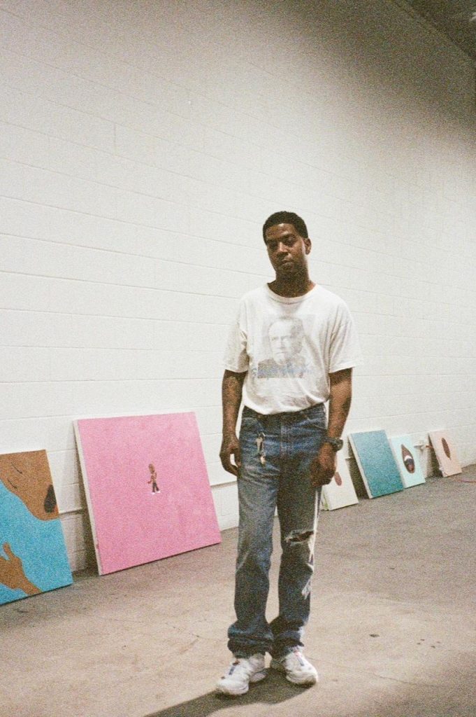 Scotty Ramon stands in a sparse studio space with several colorful canvases leaning against a white brick wall behind him.