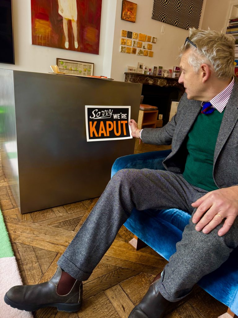 A man sitting on a blue chair inside a gallery-like room holds a black sign that reads “Sorry We’re Kaput.” Abstract paintings and small gold artworks hang on the wall behind him.