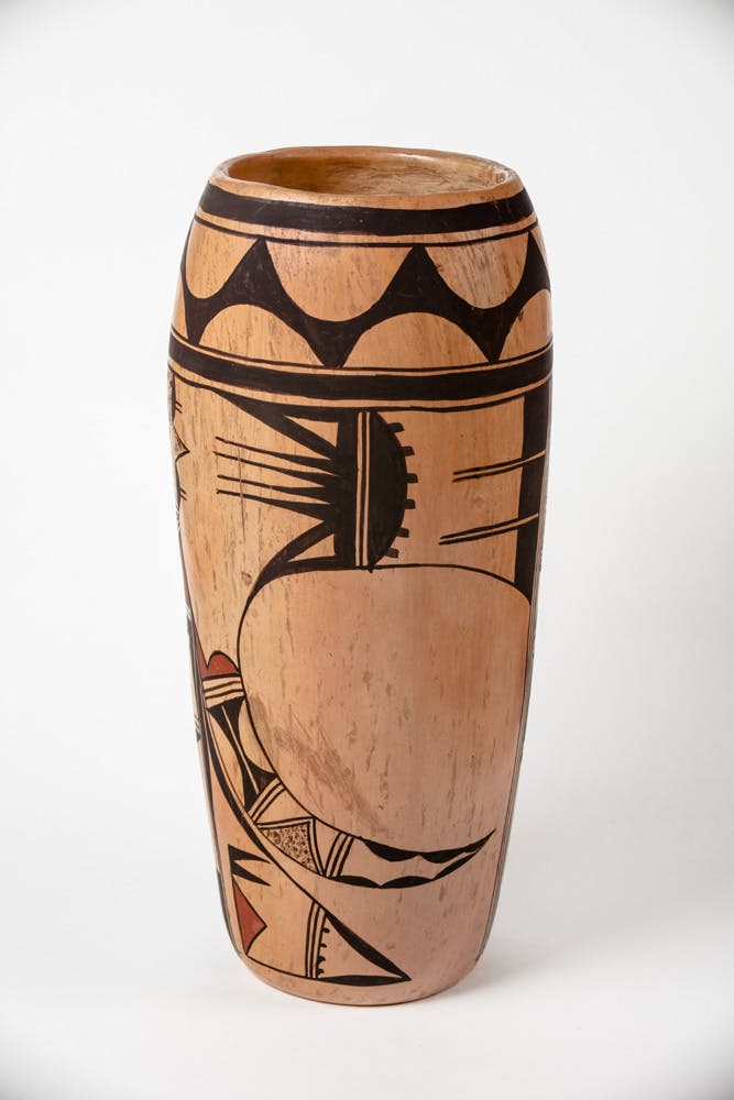 Tall Hopi-style ceramic vessel with a natural clay background and black geometric and symbolic designs, including triangular bands around the rim and a large stylized motif on the body; the vase has a rounded base, slightly tapered body, and wide opening.