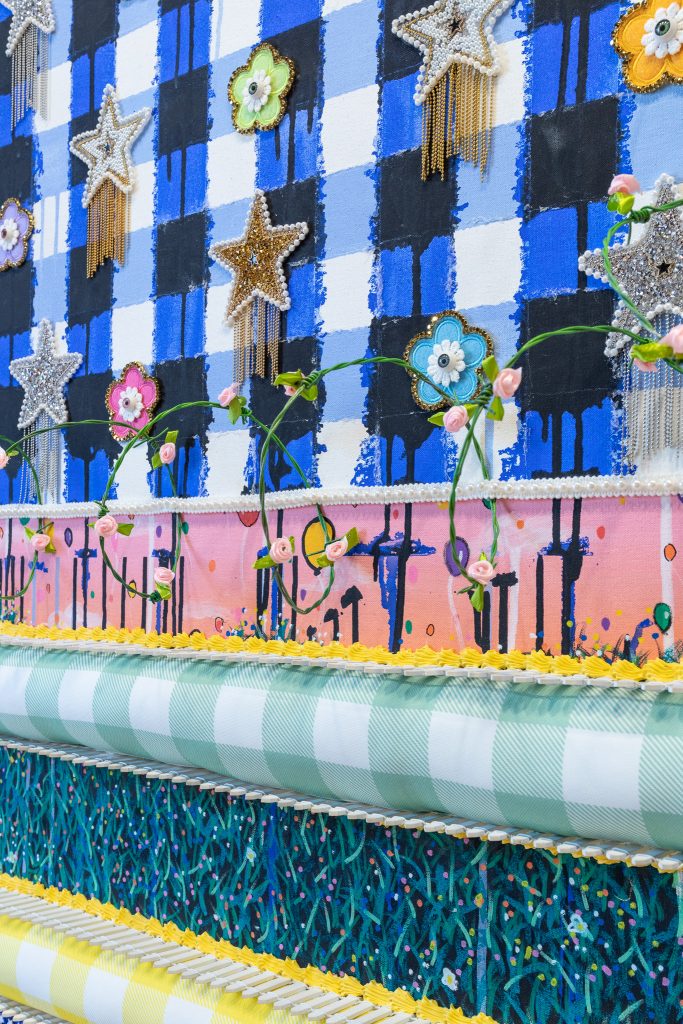 colorful mixed-media artwork featuring layered blue checkered patterns, glittery star and flower embellishments, and small pink roses arranged across textured bands.