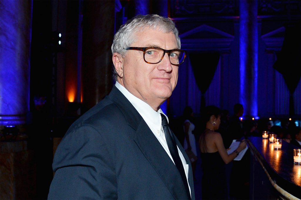 A man with glasses and grey hair wears a suit in a dark setting