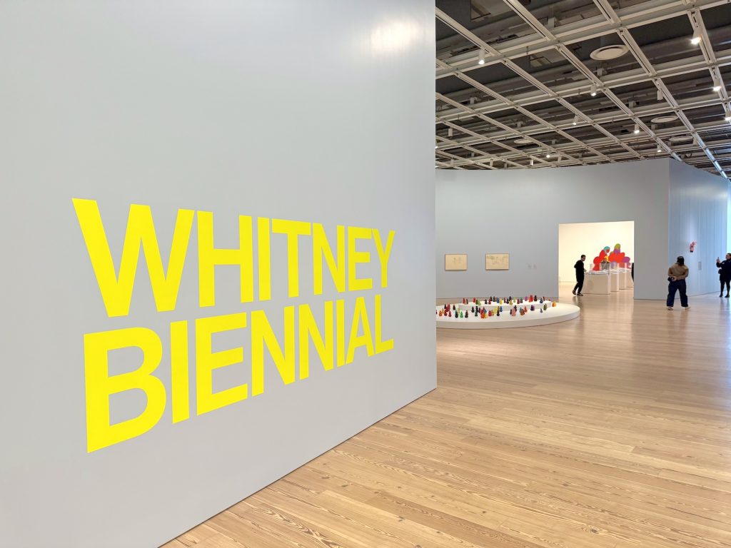 An installation view of the Whitney Biennial featuring large yellow wall text, hardwood floors, and various sculptures and artworks displayed throughout a bright gallery space.
