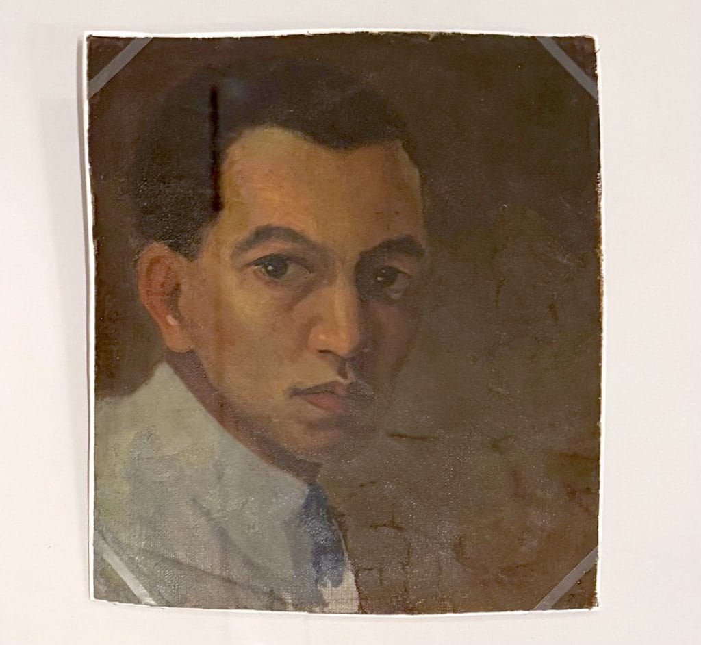 A classic oil-on-canvas self-portrait of artist Wifredo Lam, showing him from the shoulders up in a three-quarter view against a dark, muted background. He has a serious expression, dark hair, and is wearing a light-colored collared shirt with a hint of a blue tie.
