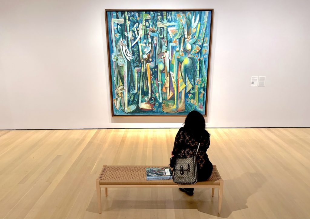 A person sits on a wooden bench viewing Wifredo Lam's "The Jungle" painting at MoMA, a large modernist work featuring abstracted figures in blues and greens with vertical bamboo-like forms.
