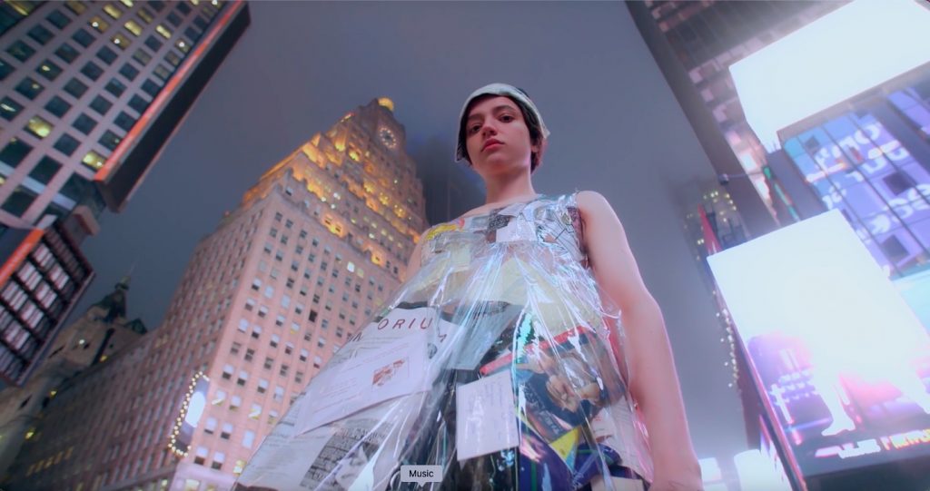 Low-angle shot of a person in a dress made of clear plastic and newspaper clippings, standing in Times Square surrounded by glowing city skyscrapers.