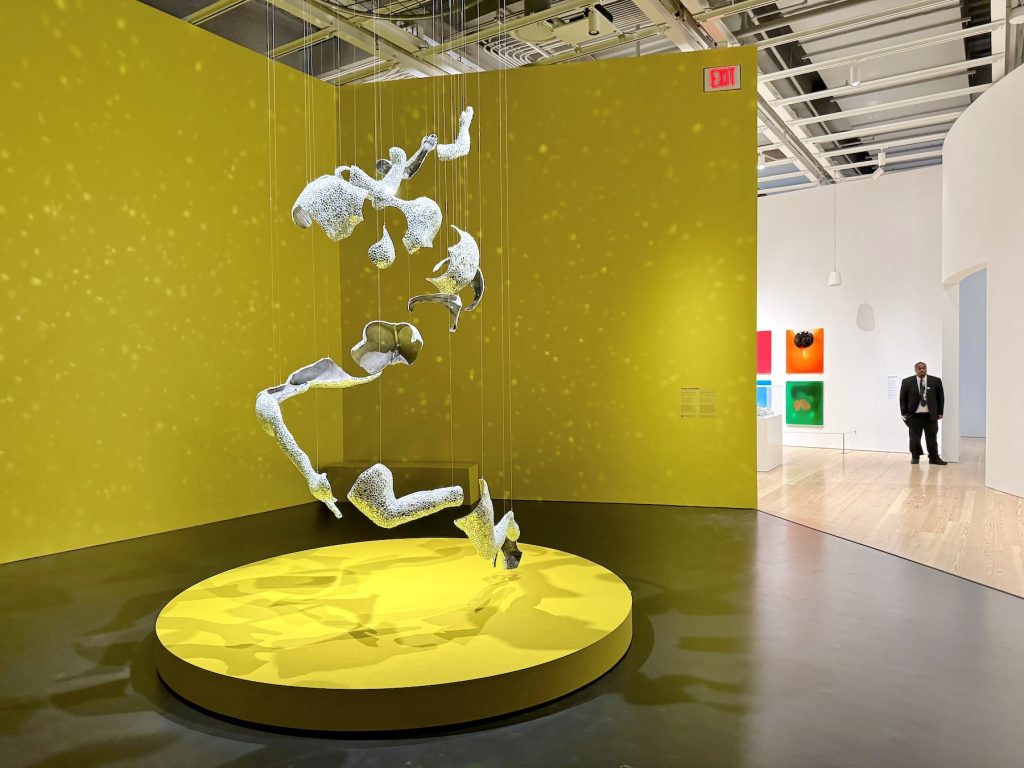 A hanging sculpture of fragmented, shimmering body parts is suspended over a circular yellow platform, set against a bright yellow corner in a modern gallery.