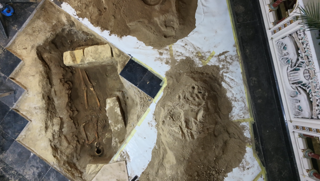 An overhead view of a hole dug into a church floor featuring the burial of a half-unearthed skeleton