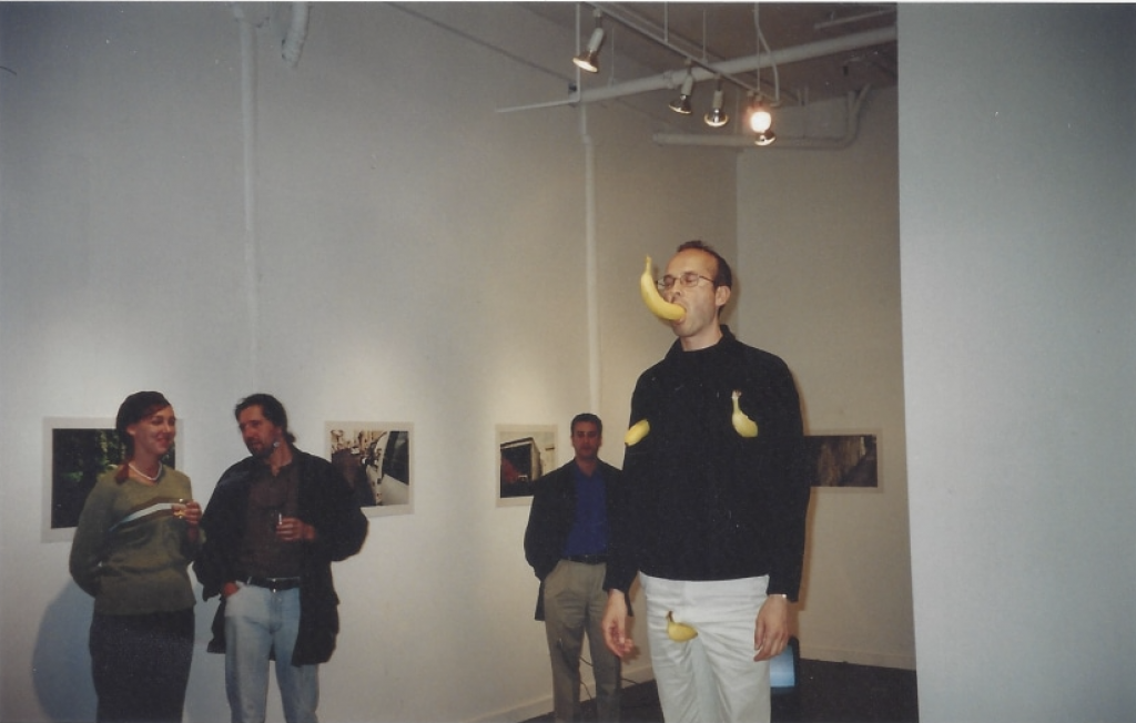 A man stands with a banana in his mouth and two bananas attached to his shirt in a dimly lit art gallery