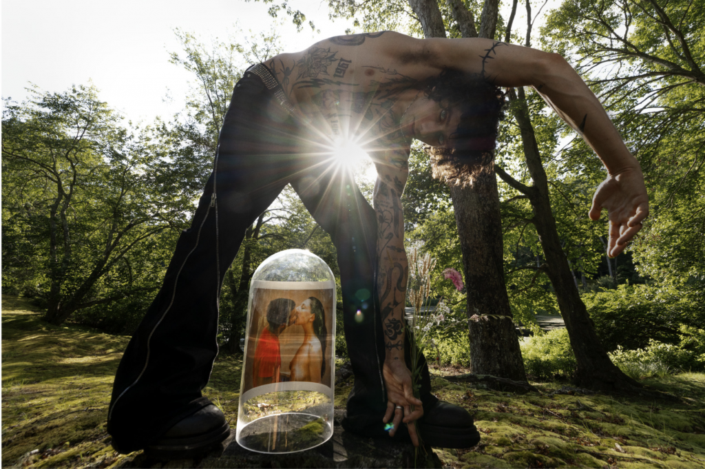 A photograph of a young, shirtless, tattoed man crouching over a cloche containing a photograph of Inez & Vinoodh kissing in a green clearing, as a bright ray of light peeks through the contours of his body