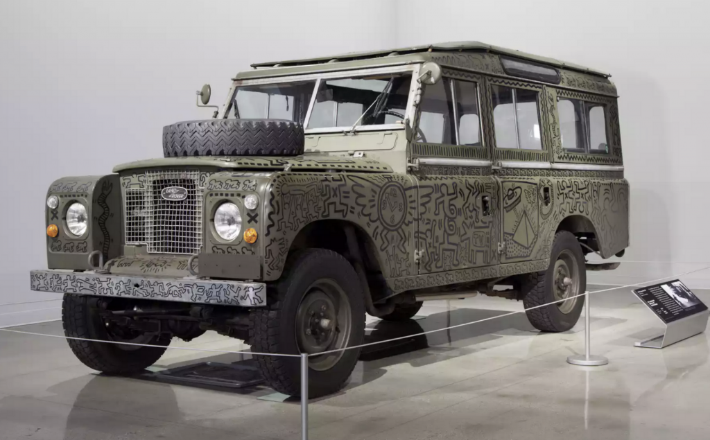 A photograph of an army green Land Rover covered in black squiggly drawings in a gray-walled gallery, with stanchions around it