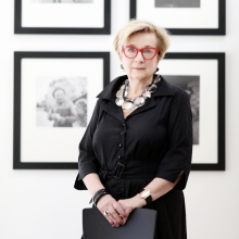 A photograph of Lynda Roscoe Hartigan standing in a simple black dress and statement necklace with very large clear beads before a white wall hung with four framed black and white photographs