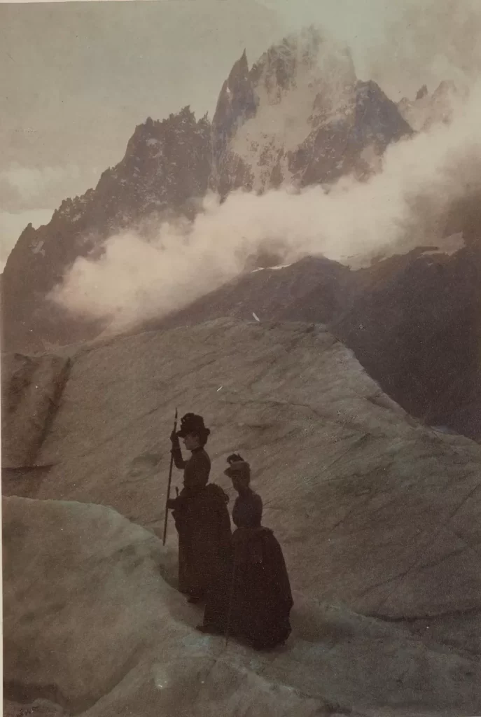 A sepia-toned photograph of two women in long dresses standing on a rocky slope beneath jagged mountain peaks partially obscured by mist.