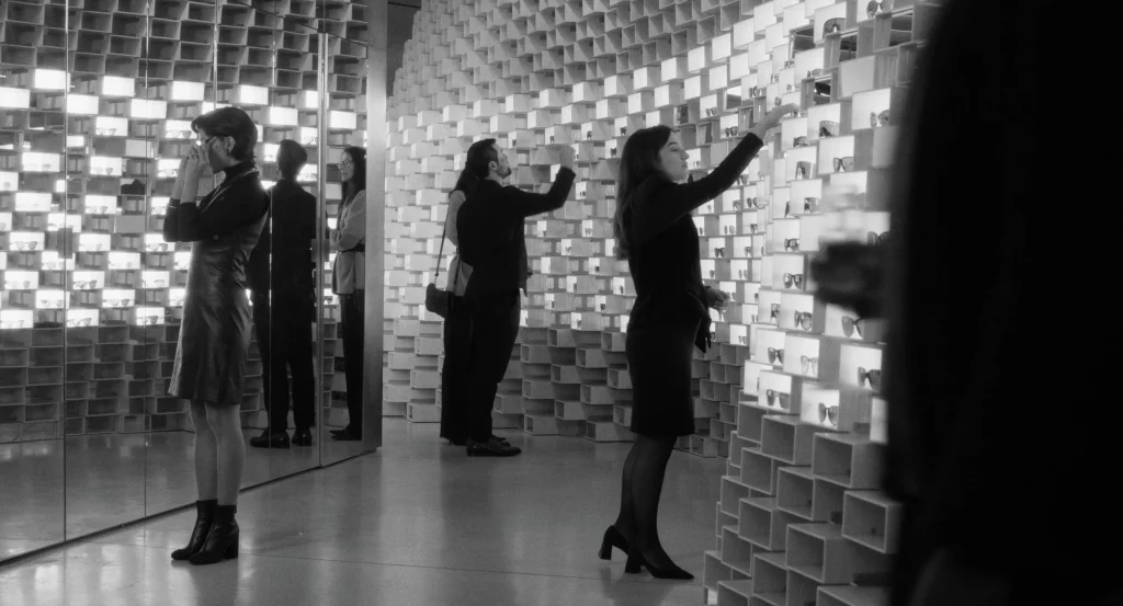 Still from Two People Exchanging saliva showing a black-and-white interior of a modern eyewear boutique, with customers examining rows of glasses displayed in illuminated grid-like shelves reflected in mirrors.